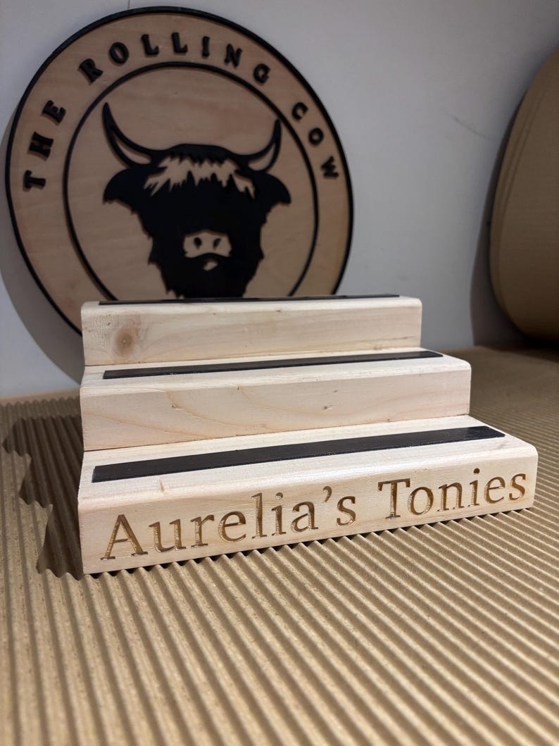 Pu&ograve; includere: Un espositore in legno a tre livelli con dettagli neri e la scritta incisa "Aurelia's Tonies". Un cartello in legno con "THE ROLLING COW" e una silhouette di mucca delle Highlands sullo sfondo.