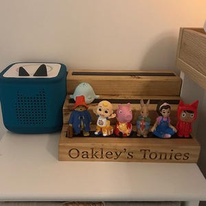 Pu&ograve; includere: Un lettore audio turchese e una mensola in legno con statuette di personaggi. La mensola &egrave; incisa con "Oakley's Tonies". Le statuette includono un orso, un bambino, un maiale, un coniglio, una ragazza e un personaggio rosso.