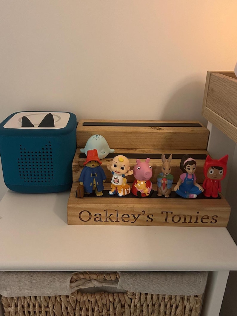 Pu&ograve; includere: Un lettore audio turchese e una mensola in legno con statuette di personaggi. La mensola &egrave; incisa con "Oakley's Tonies". Le statuette includono un orso, un bambino, un maiale, un coniglio, una ragazza e un personaggio rosso.
