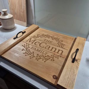 May include: A light-wood serving tray engraved with "The McCann Family" and floral designs. It features black handles and a rectangular shape. A wooden chopping board and a ceramic jar are visible in the background.