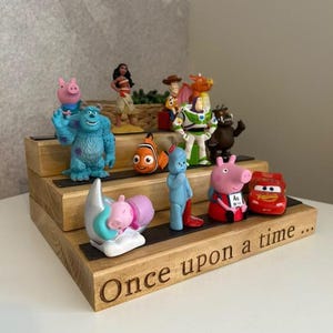 Pu&ograve; includere: Una mensola in legno a pi&ugrave; livelli con varie statuette di personaggi, tra cui Sully, Moana, Woody, Buzz Lightyear e Peppa Pig. La mensola presenta la scritta "Once upon a time..." incisa sul davanti.