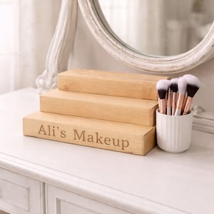 May include: A wooden makeup organiser with the text "Ali's Makeup" engraved on the front. The organiser has three stacked levels and is next to a white ceramic cup holding makeup brushes. A white framed mirror is in the background.