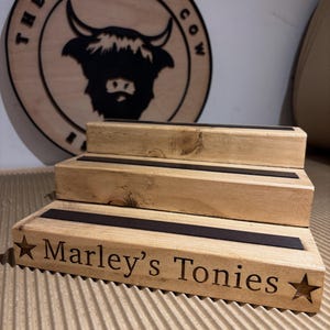 Pu&ograve; includere: Un espositore in legno a pi&ugrave; livelli con la scritta "Marley's Tonies" e una stella. L'espositore ha tre livelli con strisce nere. Un cartello in legno con il logo "The Rolling Cow" &egrave; sullo sfondo.
