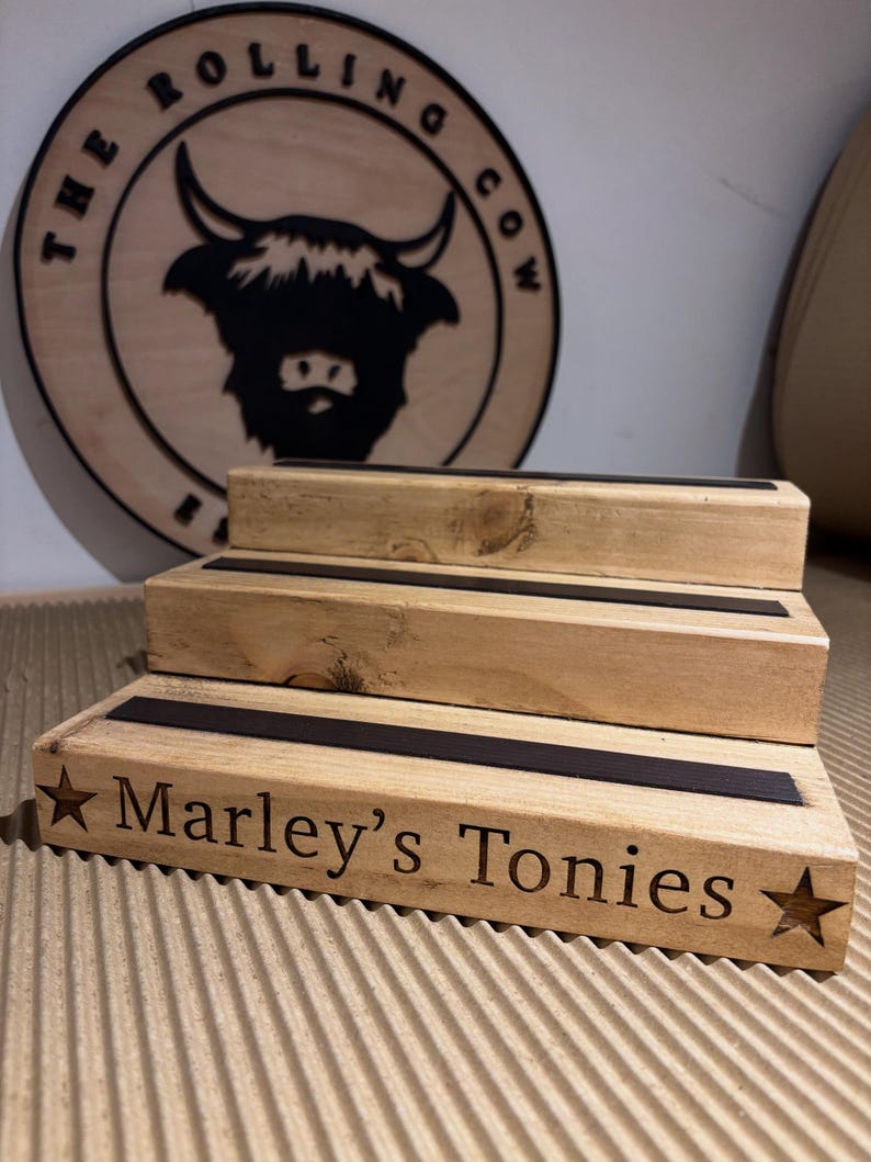 Pu&ograve; includere: Un espositore in legno a pi&ugrave; livelli con la scritta "Marley's Tonies" e una stella. L'espositore ha tre livelli con strisce nere. Un cartello in legno con il logo "The Rolling Cow" &egrave; sullo sfondo.