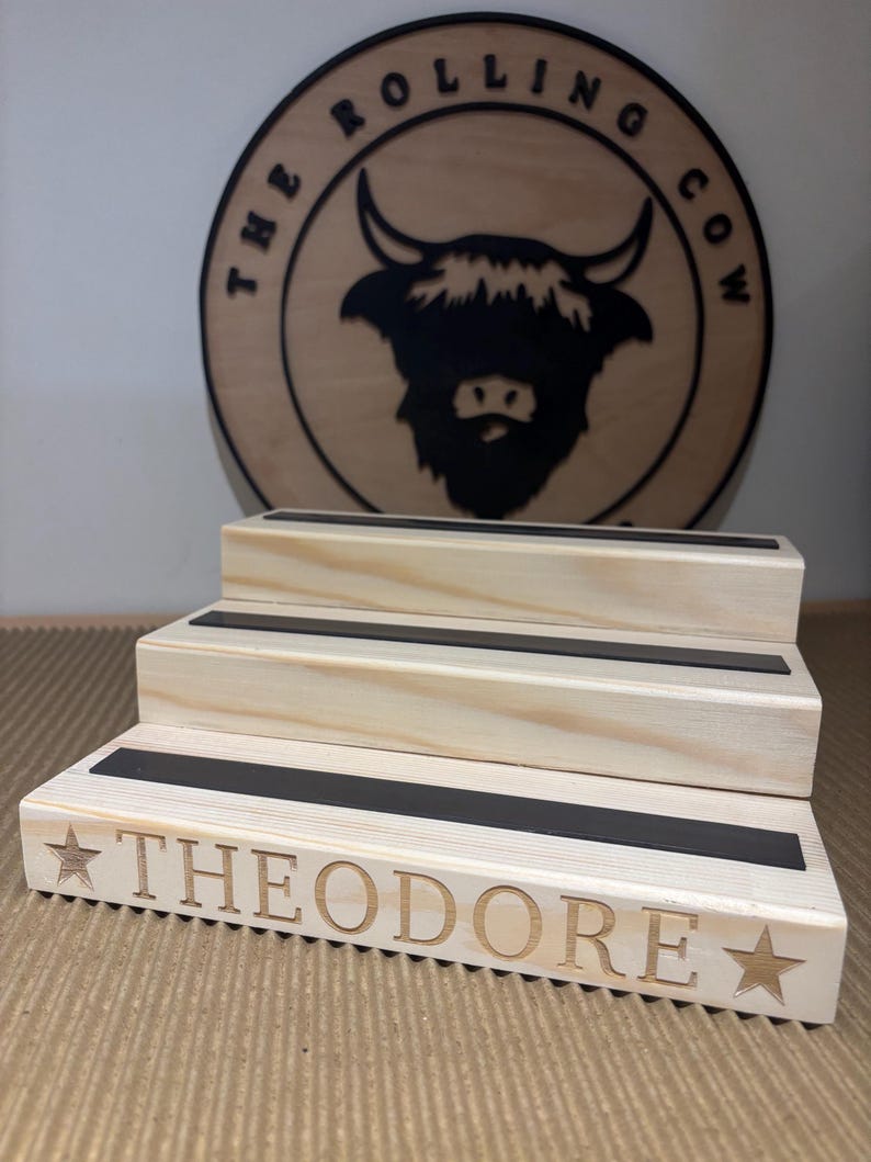 Pu&ograve; includere: Un espositore in legno a tre livelli con strisce nere. La parola "THEODORE" &egrave; incisa sul davanti, insieme a disegni di stelle. Un cartello rotondo in legno con una grafica di toro e il testo "THE ROLLING COW" &egrave; sullo sfondo.