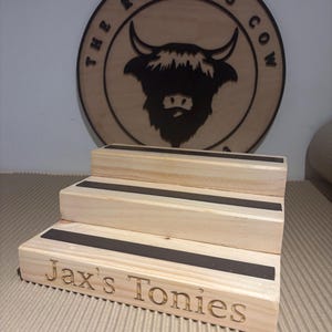 Pu&ograve; includere: Un espositore in legno a tre livelli con strisce nere. Il gradino inferiore &egrave; inciso con "Jax's Tonies". Sullo sfondo un cartello circolare in legno con "The Rolling Cow" e una silhouette di toro.