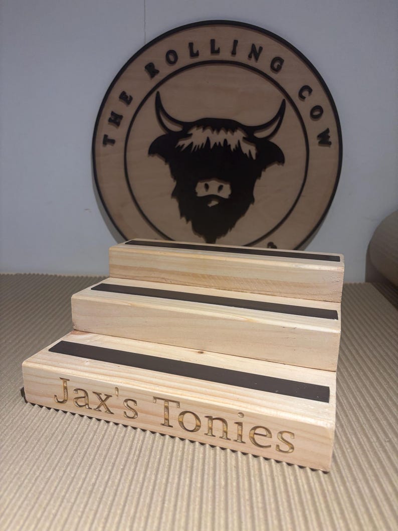Pu&ograve; includere: Un espositore in legno a tre livelli con strisce nere. Il gradino inferiore &egrave; inciso con "Jax's Tonies". Sullo sfondo un cartello circolare in legno con "The Rolling Cow" e una silhouette di toro.