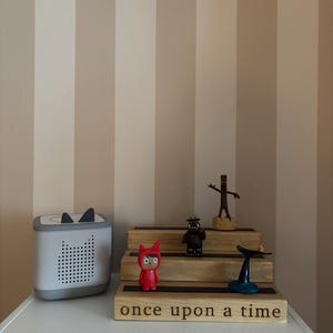 Pu&ograve; includere: Un altoparlante bianco e grigio con dettagli di orecchie di gatto nere &egrave; posto su una mensola bianca accanto a un espositore in legno con statuette. L'espositore ha le parole "once upon a time" incise.