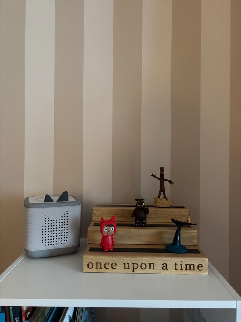 Pu&ograve; includere: Un altoparlante bianco e grigio con dettagli di orecchie di gatto nere &egrave; posto su una mensola bianca accanto a un espositore in legno con statuette. L'espositore ha le parole "once upon a time" incise.