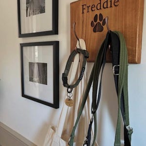 May include: A wooden dog leash holder with the name "Freddie" and a paw print. It holds a green dog collar, a cream tote bag, and green and black leads. Two framed black and white photos hang on the wall.