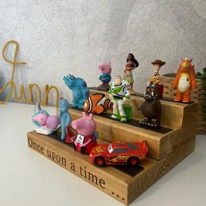 Pu&ograve; includere: Scaffale in legno a tre livelli con statuette di personaggi dei cartoni animati. Lo scaffale presenta la scritta "Once upon a time". I personaggi includono Buzz Lightyear, Woody e Saetta McQueen.