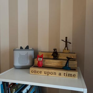 Pu&ograve; includere: Una mensola bianca con una collezione di oggetti per bambini. Un altoparlante grigio con orecchie da gatto &egrave; accanto a un espositore in legno con statuette e la scritta "once upon a time". I libri sono visibili sullo scaffale sottostante.