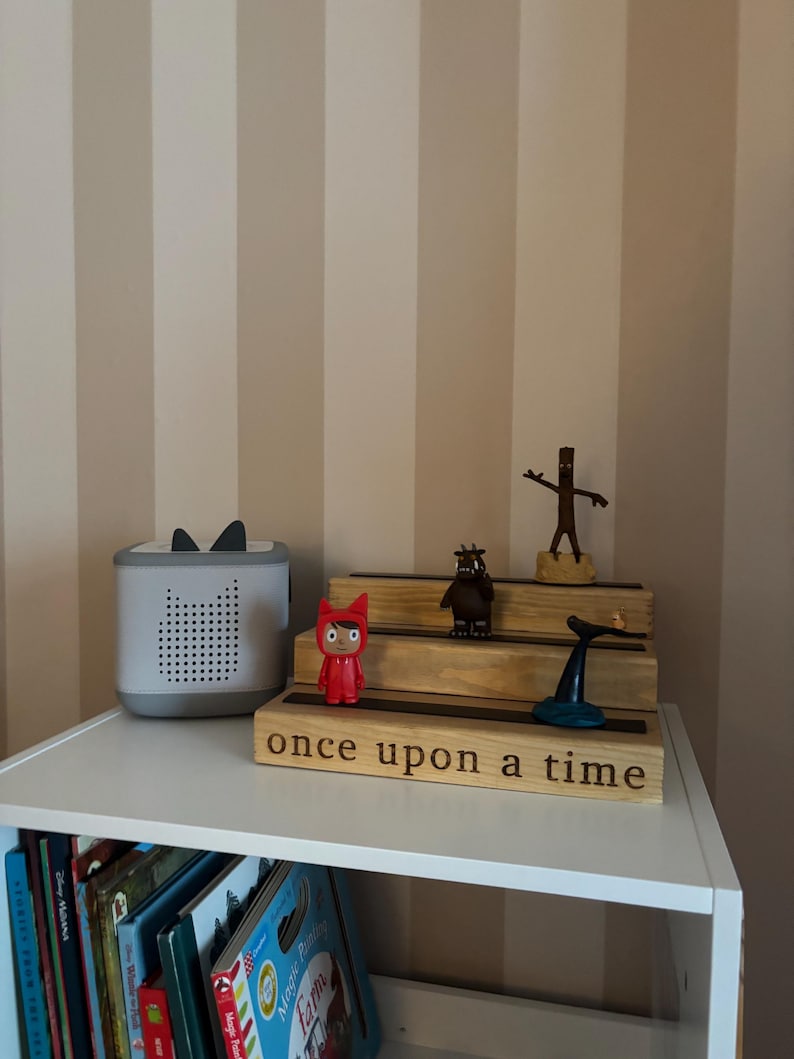 Pu&ograve; includere: Una mensola bianca con una collezione di oggetti per bambini. Un altoparlante grigio con orecchie da gatto &egrave; accanto a un espositore in legno con statuette e la scritta "once upon a time". I libri sono visibili sullo scaffale sottostante.