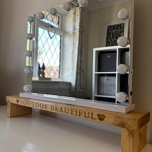 May include: A rectangular vanity mirror with a white frame and bright bulbs around the edge. It rests on a wooden base with the engraved text "YOU LOOK BEAUTIFUL" and a heart symbol. The mirror reflects a window.