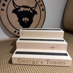 Pu&ograve; includere: Un espositore in legno a tre livelli con strisce nere su ogni gradino. Il gradino inferiore &egrave; inciso con "George's Tonies". Sullo sfondo un cartello in legno con "The Rolling Cow" e una silhouette di mucca delle Highlands.