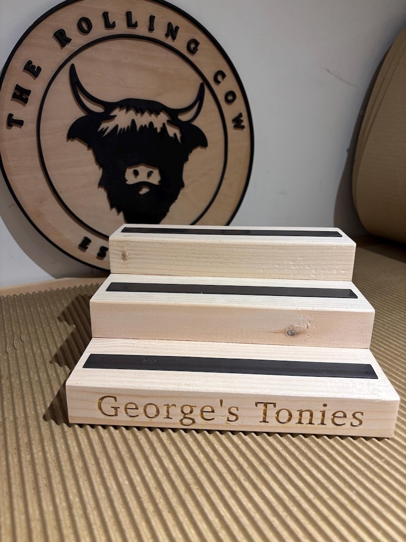 Pu&ograve; includere: Un espositore in legno a tre livelli con strisce nere su ogni gradino. Il gradino inferiore &egrave; inciso con "George's Tonies". Sullo sfondo un cartello in legno con "The Rolling Cow" e una silhouette di mucca delle Highlands.
