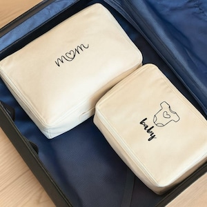 May include: Two off-white travel organizers inside a blue suitcase. One organizer has the word "mom" with a heart, and the other has the word "baby" with a baby onesie graphic. Both have a zipper closure.