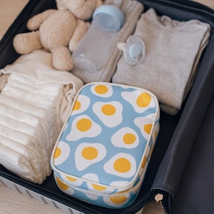 May include: A suitcase filled with baby items. A blue cosmetic bag with a fried egg pattern is visible. Other contents include a teddy bear, nappies, a baby bottle, a grey sweater, and a dummy.