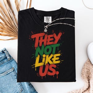 Op de afbeelding: Zwart T-shirt met de tekst "THEY NOT LIKE US" in rode, groene en gele graffiti-stijl letters. Een zilveren ketting met een zonnehanger ligt op het shirt. Het shirt is opgevouwen op een witte ondergrond met een spijkerbroek en een gedroogde plant.