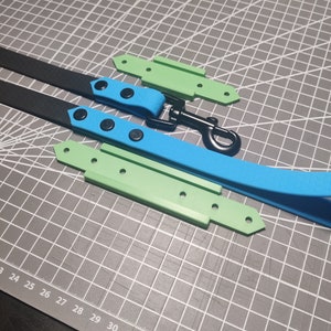 Pode incluir: Duas trelas para cães, uma preta e uma azul, com ferragens de metal preto e acessórios de plástico verde. As trelas estão dispostas sobre um tapete de corte cinza.