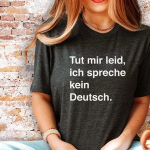 May include: Dark gray t-shirt with the white text "Tut mir leid, ich spreche kein Deutsch." The shirt has a crew neck and short sleeves. The person is wearing a gold necklace and bracelets.