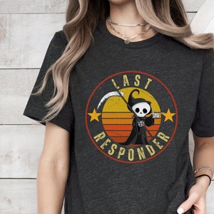 May include: A dark gray t-shirt with a retro-style graphic of a cartoon grim reaper holding a scythe. The graphic is surrounded by a circle with the text "Last Responder" in a vintage font.