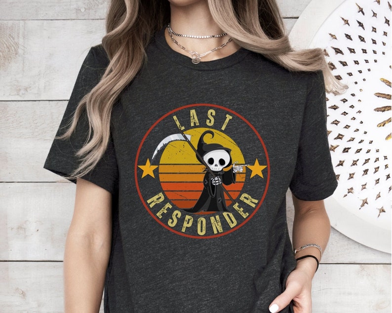 May include: A dark gray t-shirt with a retro-style graphic of a cartoon grim reaper holding a scythe. The graphic is surrounded by a circle with the text "Last Responder" in a vintage font.