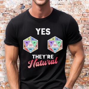 May include: Black t-shirt with the text "YES THEY'RE Natural" and two colorful D20 dice graphics. The dice are multi-sided with numbers visible. The shirt is a crew neck style.