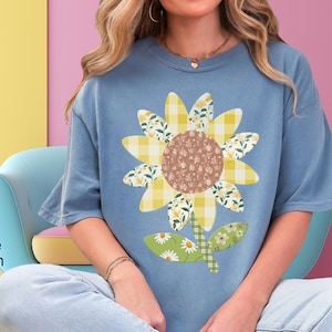 May include: Blue jean-colored t-shirt featuring a large sunflower design. The sunflower has a brown center, yellow gingham and floral patterned petals, and a green stem with a daisy and floral pattern. The text "Blue Jean" is on the left side.