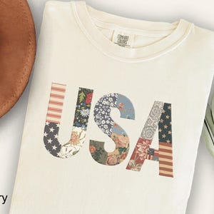 May include: An ivory-colored t-shirt featuring the letters USA printed in a patchwork style. The letters are filled with various patterns including red and white stripes, blue with white stars, and floral designs in muted tones of red, blue, and green.
