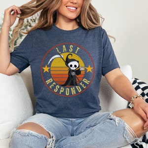 May include: A navy blue t-shirt with a retro-style graphic of a cartoon Grim Reaper holding a drink. The graphic is surrounded by a circle with the text "Last Responder" in a vintage font.