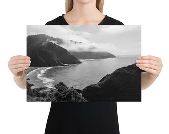 Op de afbeelding: Een zwart-witfoto van een kustlandschap met een rotsachtige kustlijn, een baai met water en een beboste heuvel op de achtergrond. Het water is kalm en de lucht is bewolkt.