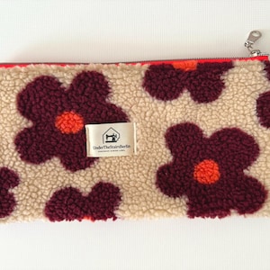 May include: A rectangular, beige fleece pouch with a repeating pattern of large, burgundy flowers with orange centers. The pouch has a red zipper and a silver zipper pull. A small tag reads "UnderTheStairsBerlin".