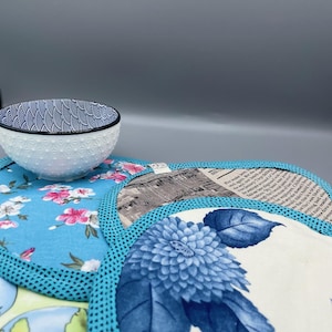 Peut inclure: Un bol blanc et bleu à motifs est posé sur une collection de sets de table ronds en tissu. Les sets de table présentent des motifs floraux et de cartes du monde, avec une bordure bleue. Un set de table affiche une grande impression florale bleue.