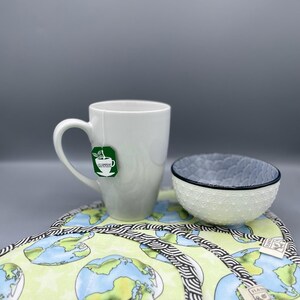 Könnte beinhalten: Eine weiße Keramiktasse mit einem Clipper-Teebeutel, eine kleine weiße Schüssel mit strukturiertem Design und eine grün-blaue Tischset mit Erdmotiv. Das Tischset hat einen schwarz-weißen Rand.