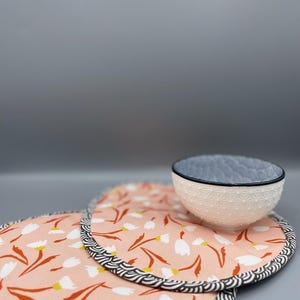 Könnte beinhalten: Eine weiße Schüssel mit strukturierter Oberfläche und blauem Rand steht auf zwei runden, rosa Tischsets. Die Tischsets zeigen ein Blumenmuster mit weißen Blüten und braunen Stielen sowie einen schwarz-weißen Wellenrand.