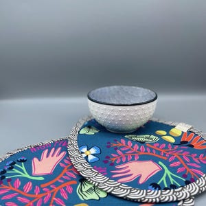 Könnte beinhalten: Eine kleine weiße Schüssel mit schwarzem Rand steht auf zwei blauen Tischsets. Die Tischsets haben ein farbenfrohes Blumendesign mit rosa, orangefarbenen und gelben Akzenten. Die Tischsets haben einen dekorativen Rand.
