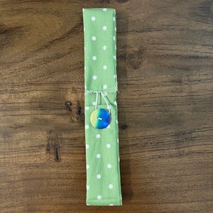 May include: A green fabric item with white polka dots, folded and secured with a button. The button is round with a gradient of blue, green, and yellow. The item appears to be a utensil holder.