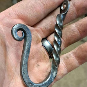 May include: Hand-forged metal hook with a twisted stem and a looped end. The hook is a dark gray color and appears to be made of iron. The hook has a hole at the top for hanging. The hook is held in a hand.