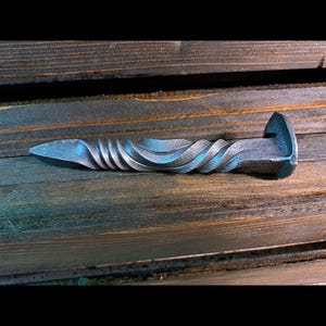May include: A hand-forged metal railroad spike with a twisted shaft and a pointed end. The spike is a dark, metallic color and rests on a wooden surface. The design features a decorative, spiraled pattern.