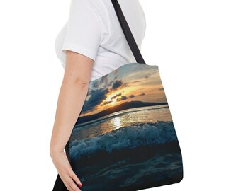 Puede incluir: Bolso tote con un vibrante atardecer sobre el océano. La imagen muestra una ola rompiendo en primer plano, con el sol poniéndose detrás de una cordillera. El bolso tiene una correa negra y se lleva al hombro.
