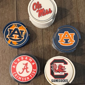 May include: A collection of five circular drink coasters featuring various collegiate sports team logos. The designs include Ole Miss, two Auburn University logos, Alabama Crimson Tide, and South Carolina Gamecocks, displayed on white and blue bases.