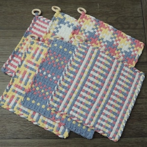 May include: Set of four woven kitchen dishcloths in blue, pink, yellow, and white. The cloths are made with a waffle weave pattern and have wooden rings attached to the corners.