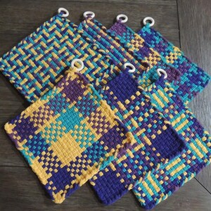 May include: Set of six woven pot holders in a purple, teal, and yellow plaid pattern. Each pot holder has a white ring attached to the top corner.