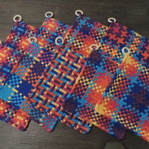 May include: A collection of colorful woven potholders. Each square potholder features a vibrant pattern of interwoven threads in shades of blue, orange, yellow, and purple. White loops are attached to the corners for hanging. The potholders are arranged on a wooden surface.