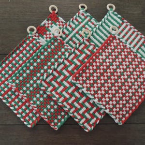 May include: Six woven potholders in red, green, and white, with a wooden ring for hanging. The potholders feature a woven pattern of red, green, and white, with a square shape. The potholders are arranged on a dark wooden surface.