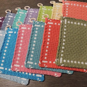 May include: A collection of colorful woven potholders in various shades, including blue, pink, green, and orange. Each potholder has a white dotted border and a wooden ring for hanging. The potholders are rectangular and appear to be handmade.