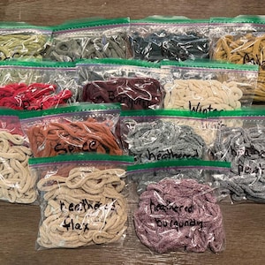Könnte beinhalten: Eine Sammlung durchsichtiger Plastikbeutel gefüllt mit verschiedenfarbigen gewickelten Schnüren oder Garnen. Jeder Beutel ist mit handschriftlichem Text beschriftet, der Farben wie flax, spice, heathered flax, heathered burgundy, red, burgundy, winter white und pewter identifiziert.