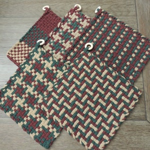 May include: Four woven pot holders in burgundy, green, and beige. Each square pot holder has a loop for hanging. The pot holders feature various woven patterns, including a herringbone and a checkerboard design.