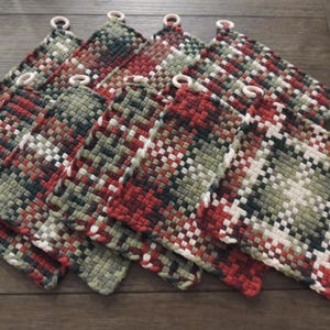 May include: A collection of woven pot holders in a plaid pattern. The pot holders feature a mix of red, green, and white threads, with wooden ring loops for hanging. The items are arranged on a wooden surface.